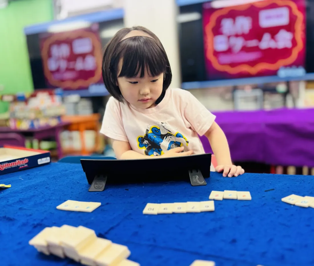 ボードゲーム「ラミィキューブ」の場に並んだ数字タイルを見つめ、次の一手を考える女の子
